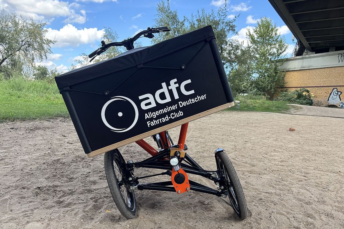 Chike schräg Ein dreirädriges Lastenrad mit schräg geneigter Transportbox steht auf einem sandigen Weg.